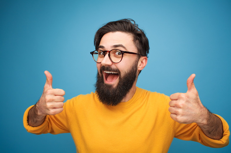 Stylish young hipster with beard wearing eyeglasses and holding thumbs up on blue.