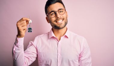 Young real estate business man holding new house keys over pink background with a happy face standing and smiling with a confident smile showing teeth